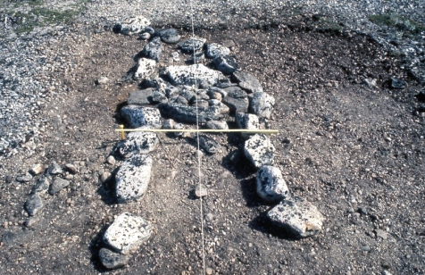 Attu's Point Locality 7 mid-passage structure with box hearth 1993. The two lines of stone forming the mid passage structure can be seen in the foreground, the box hearth is visible in the middle, just beyond the yellow ruler. The arms of the mid passage structure extend into the distance beyond the box hearth. (Hood)